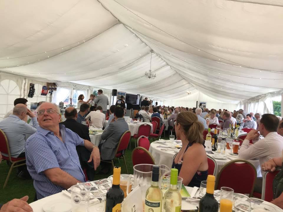 A large gathering of people seated at round tables inside a white tent during a celebration or banquet. Attendees are engaged in conversations, with some standing and near a stage at the front with speakers and a screen. The tables are decorated with drinks, bottles of wine and beer, and tableware.