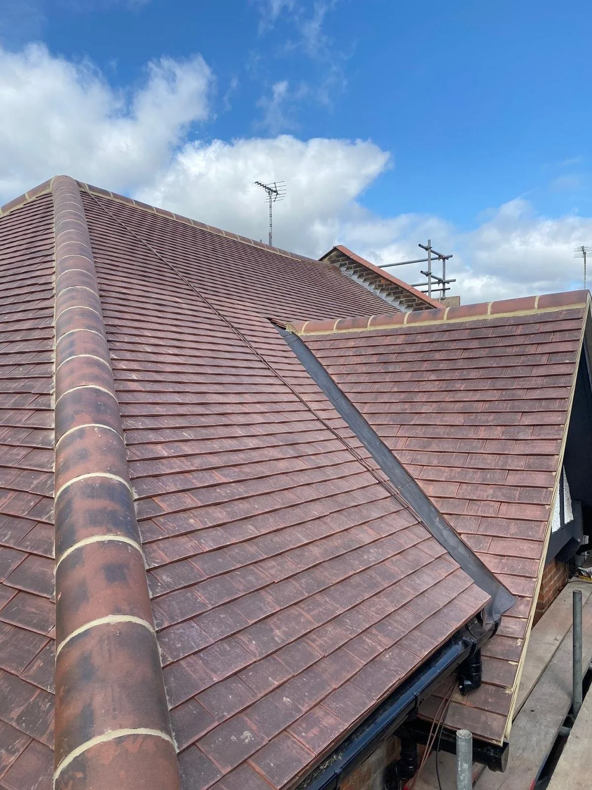 Newly fitted rooftop in north london with red clay tiles under a partly cloudy sky, with TV antennas and scaffolding visible in the background. By K G Croft.