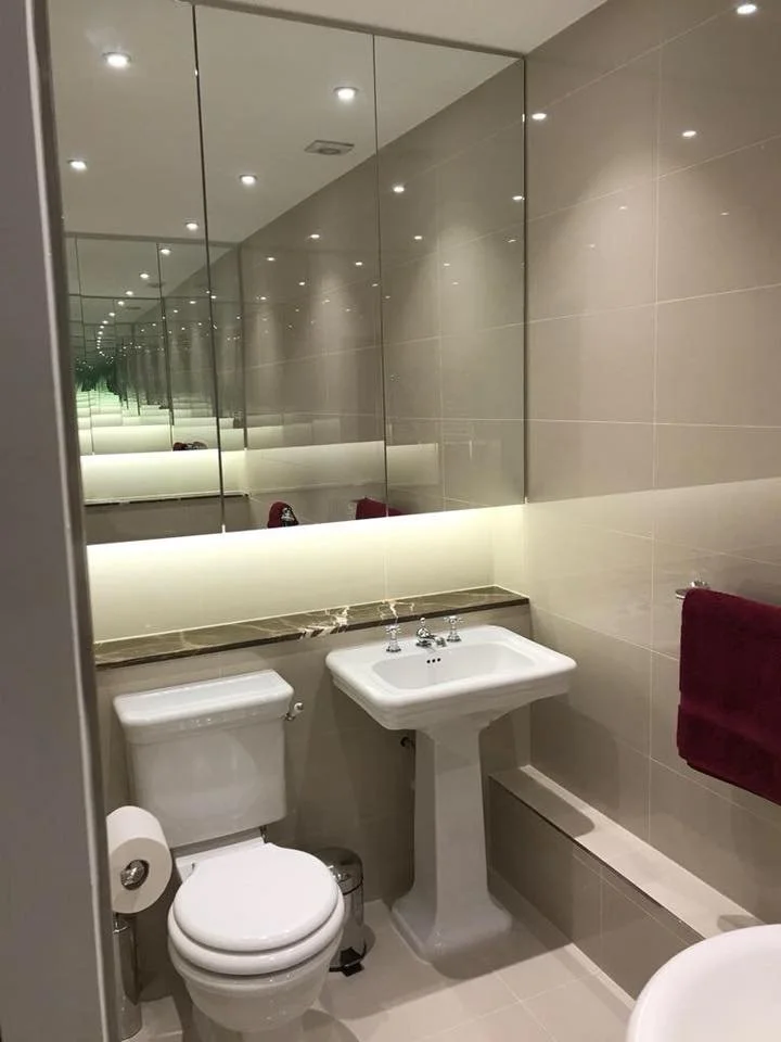 A small modern bathroom with mirrored wall panels, a toilet, a white pedestal sink, and a burgundy towel hanging on a towel rack. The bathroom features beige tiled walls and ceiling recessed lighting.