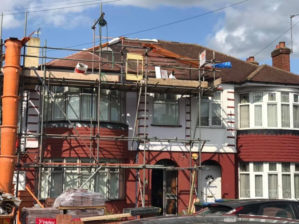 A two-story house in north london under renovation with scaffolding around the front, some construction materials, and a cloudy sky overhead. Work by K G Croft.