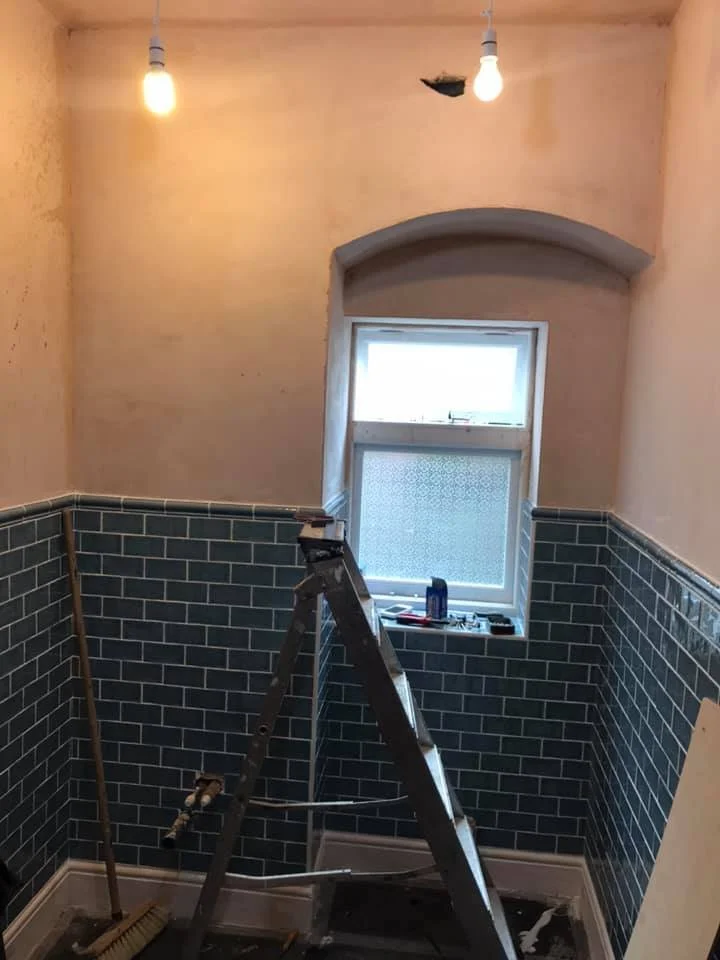 A partially renovated bathroom with two exposed light bulbs hanging from the ceiling, a small window, and black subway tiles on the lower half of the walls. There is a ladder, a broom, and construction tools visible in the room.