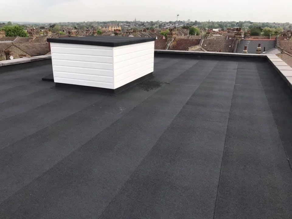 Flat rooftop by K G Croft in north london with black roofing material, a white rectangular vent or chimney with a black top, and a cityscape in the background with rooftops and trees.