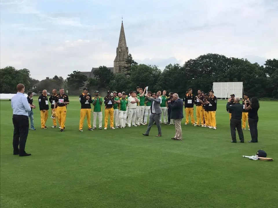 Southgate Adelaide cricket club, including athletes in yellow and black uniforms and others in casual attire, standing on a grassy field during an outdoor event or ceremony, with a church steeple and trees in the background. Sponsored by K G Croft.