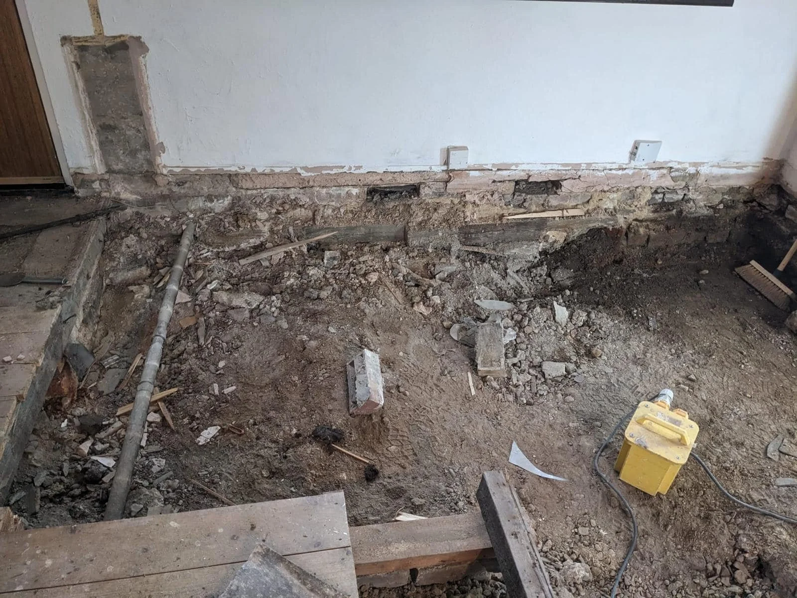 Room undergoing renovation by K G Croft in north london with exposed dirt, debris, and construction tools on the floor, and a partially removed wall with electrical outlets.