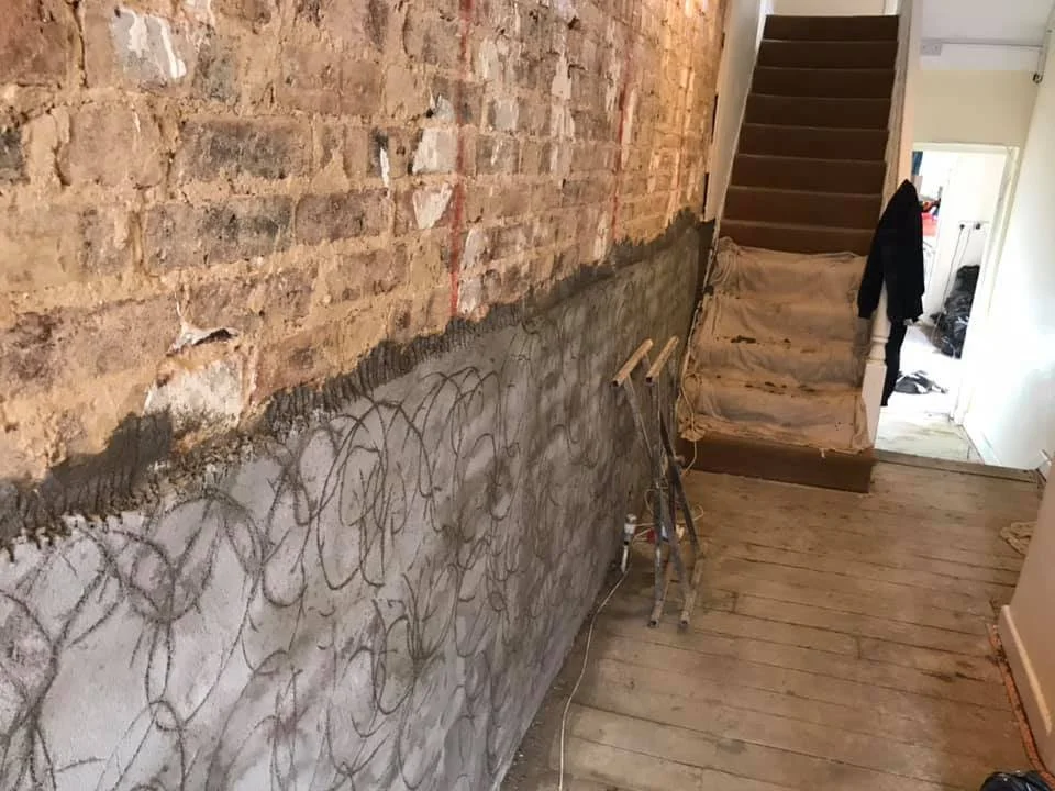 Interior of a house under renovation, showing an exposed brick wall and a section with fresh concrete or plaster with scribbled markings, a wooden staircase, and construction tools.