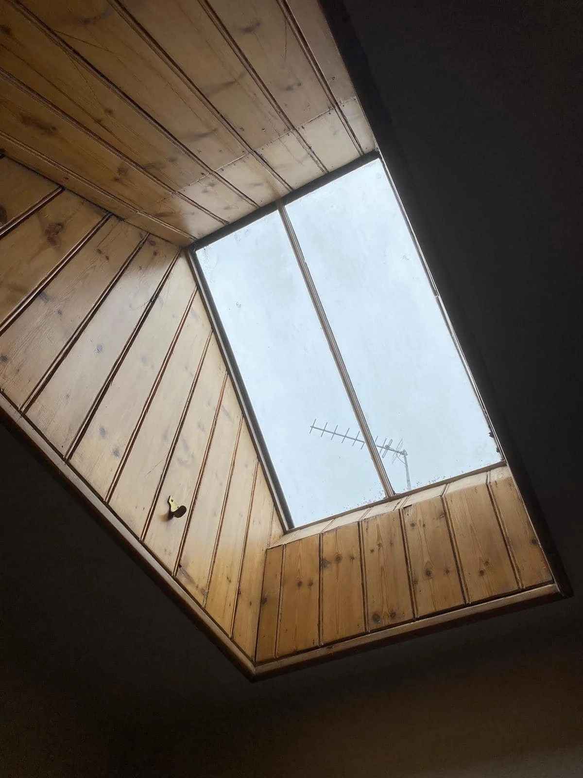 A ceiling with wood paneling surrounds a large rectangular skylight, through which a cloudy sky and TV antenna are visible. Built by K G Croft.