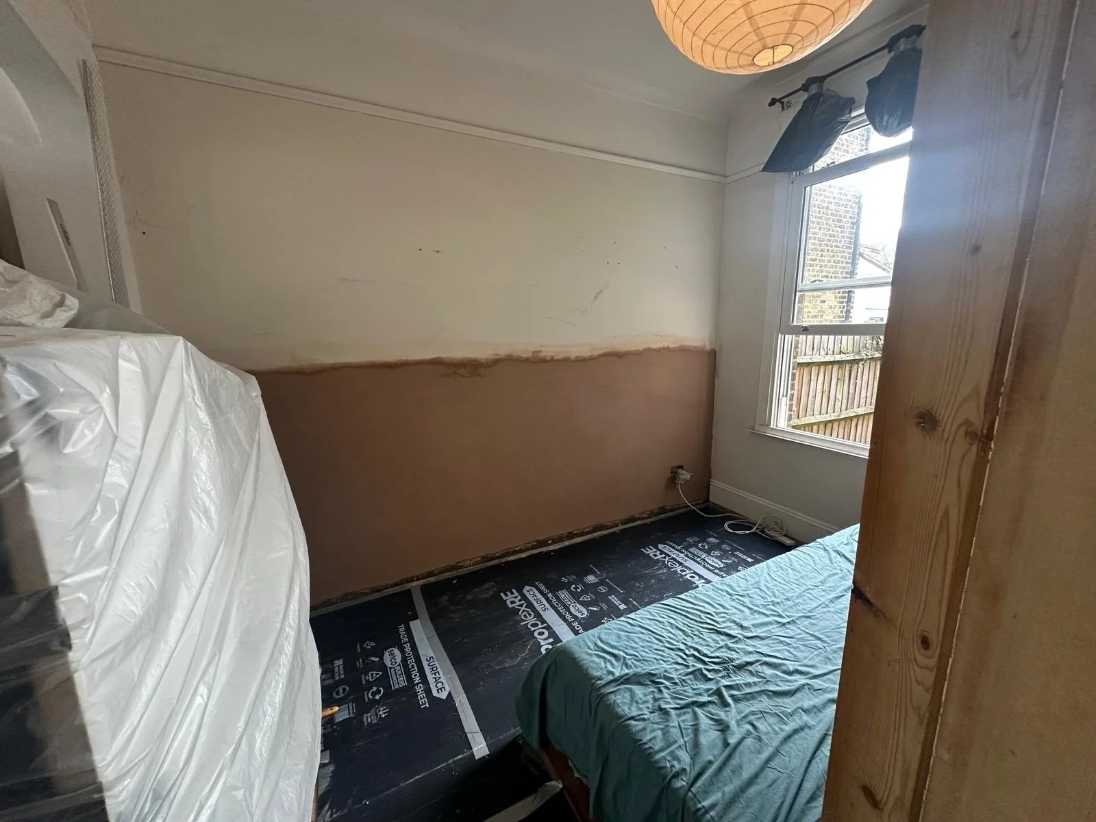 A small room under renovation with a window, partially painted wall, a bed with a green cover, and a black floor covering.