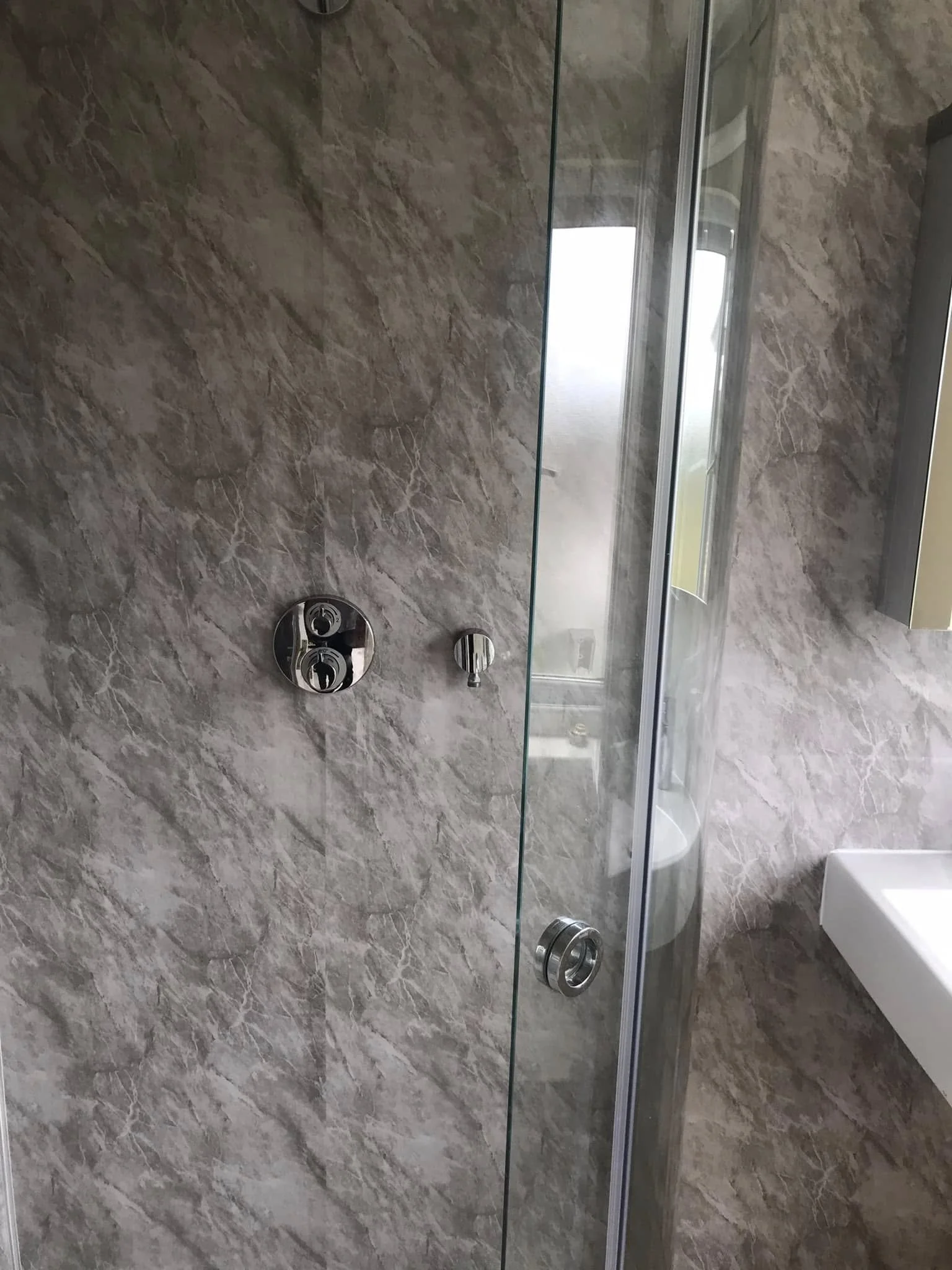 A modern bathroom shower with a glass door, marble walls, a silver shower control panel, and a partial view of a white sink.