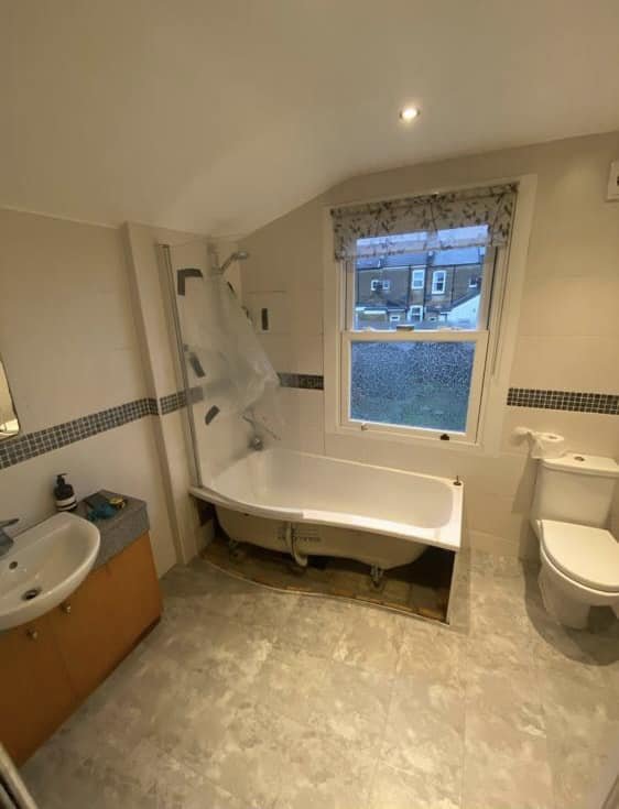 Bathroom with a bathtub, toilet, small sink, and window, area around the bathtub is under renovation with exposed plumbing.