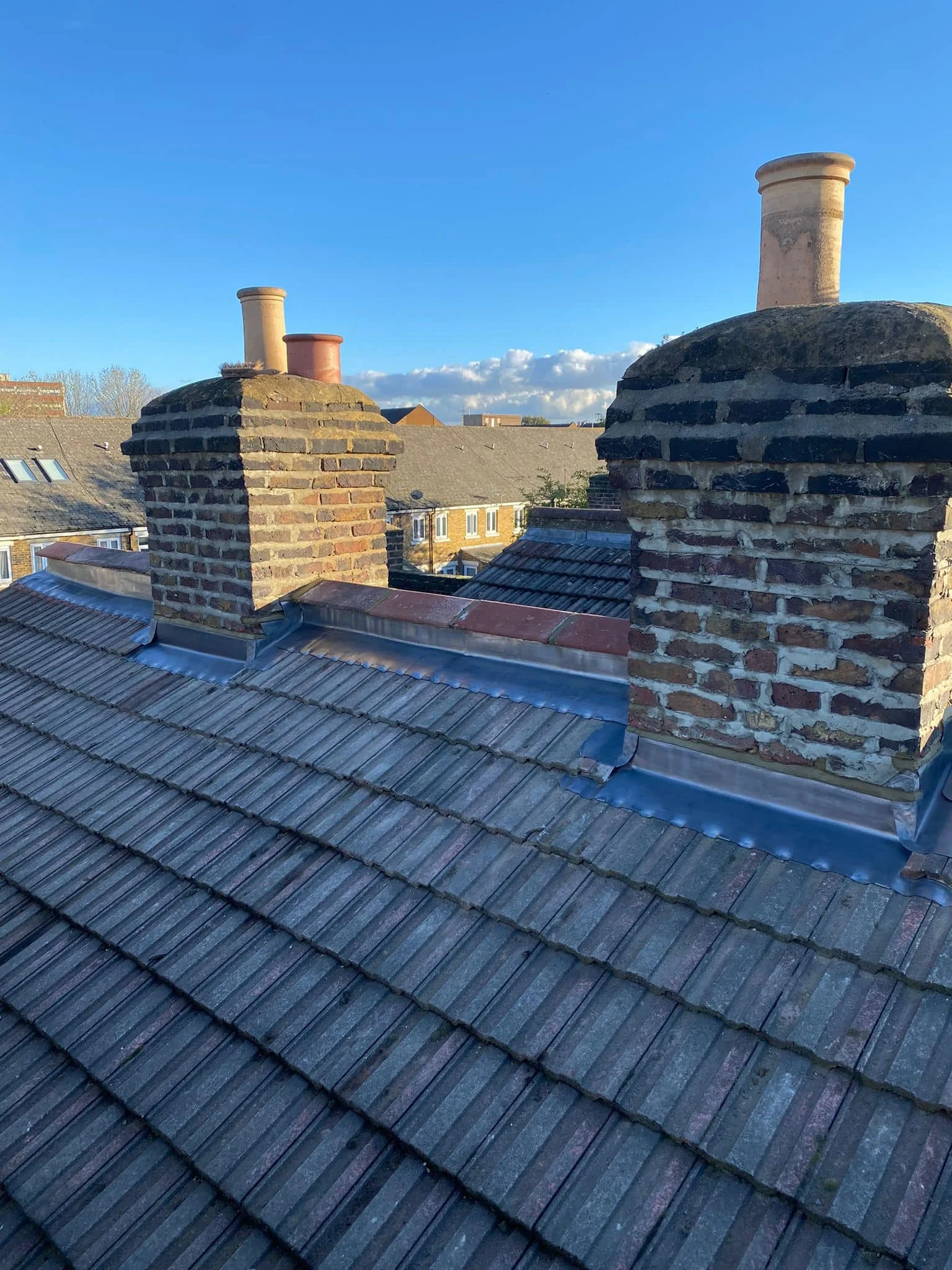 View of rooftops with brick chimneys and grey tiles, under a clear blue sky with some clouds. New roof works by K G Croft in north london.