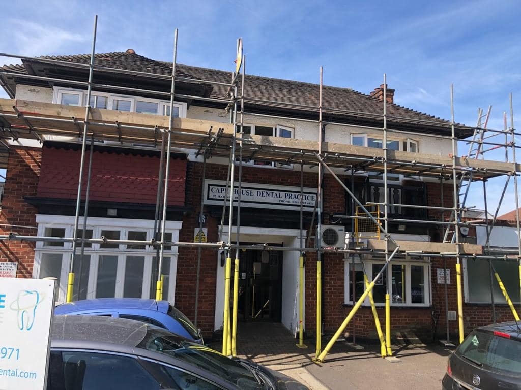 A brick building in north london with scaffolding set up around the front, indicating construction or renovation work. A sign reads "CAP House Dental Practice," and there are vehicles parked in front. K G Croft renovating.