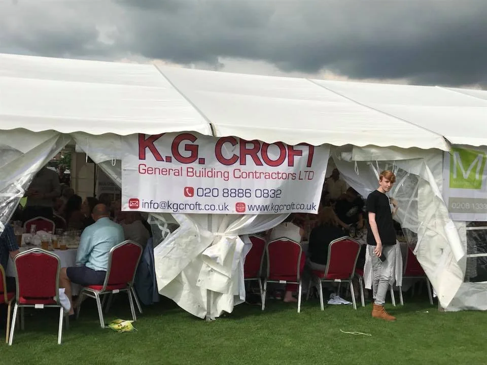 A large white event tent with a banner for K.G Croft General Building Contractors LTD hanging on it. Inside the tent are people sitting at round tables with red chairs. There is a young person standing outside the tent on the right side.