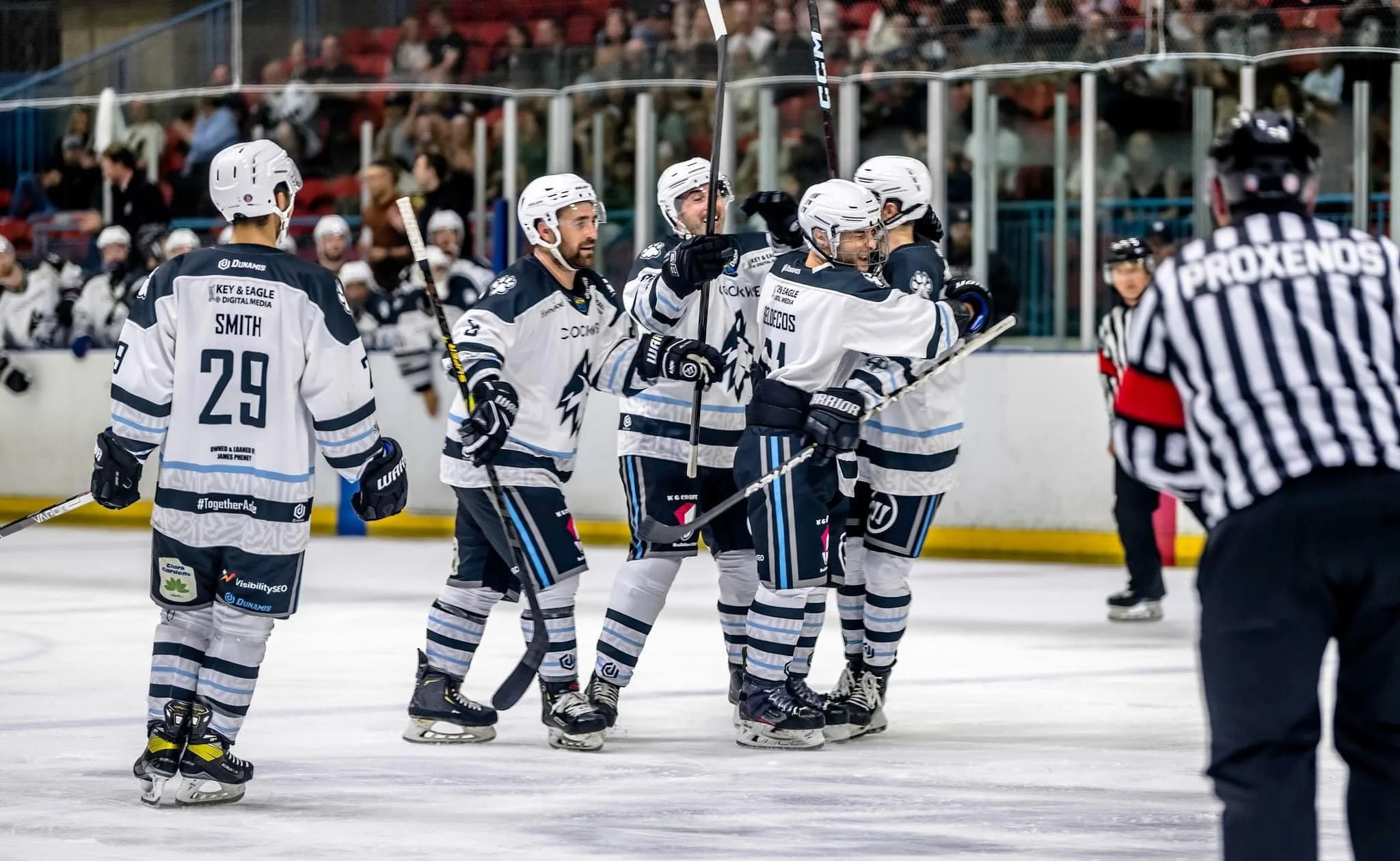 Harringay Huskies ice hockey players celebrating on the ice rink after scoring, with several teammates gathered together, and a referee in the foreground. Sponsored by K G Croft.