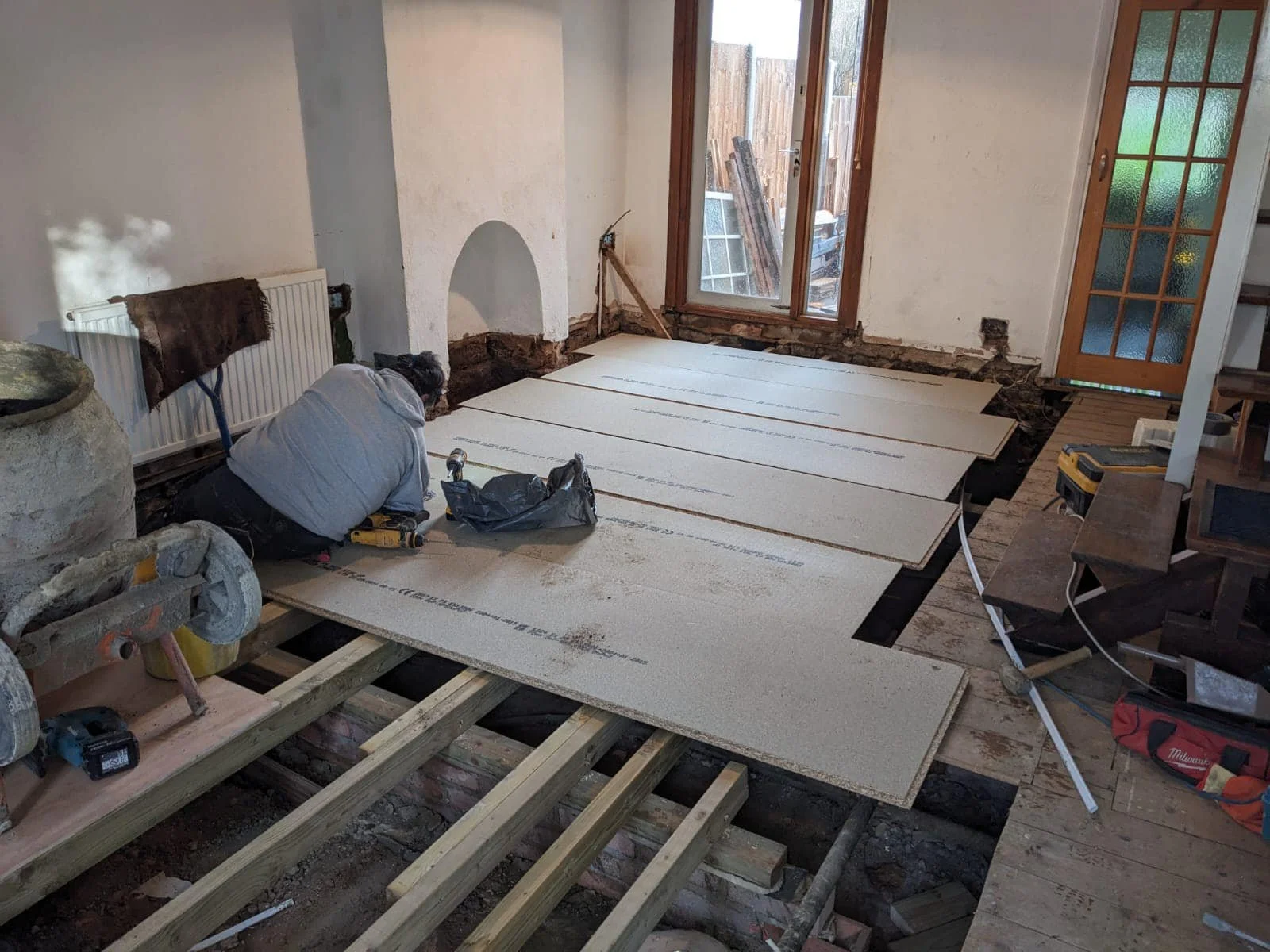K G Croft renovating a home in North london, installing flooring with large tiles or panels inside a room. The room has wooden framing on the floor, a sliding glass door, and a painted fireplace.