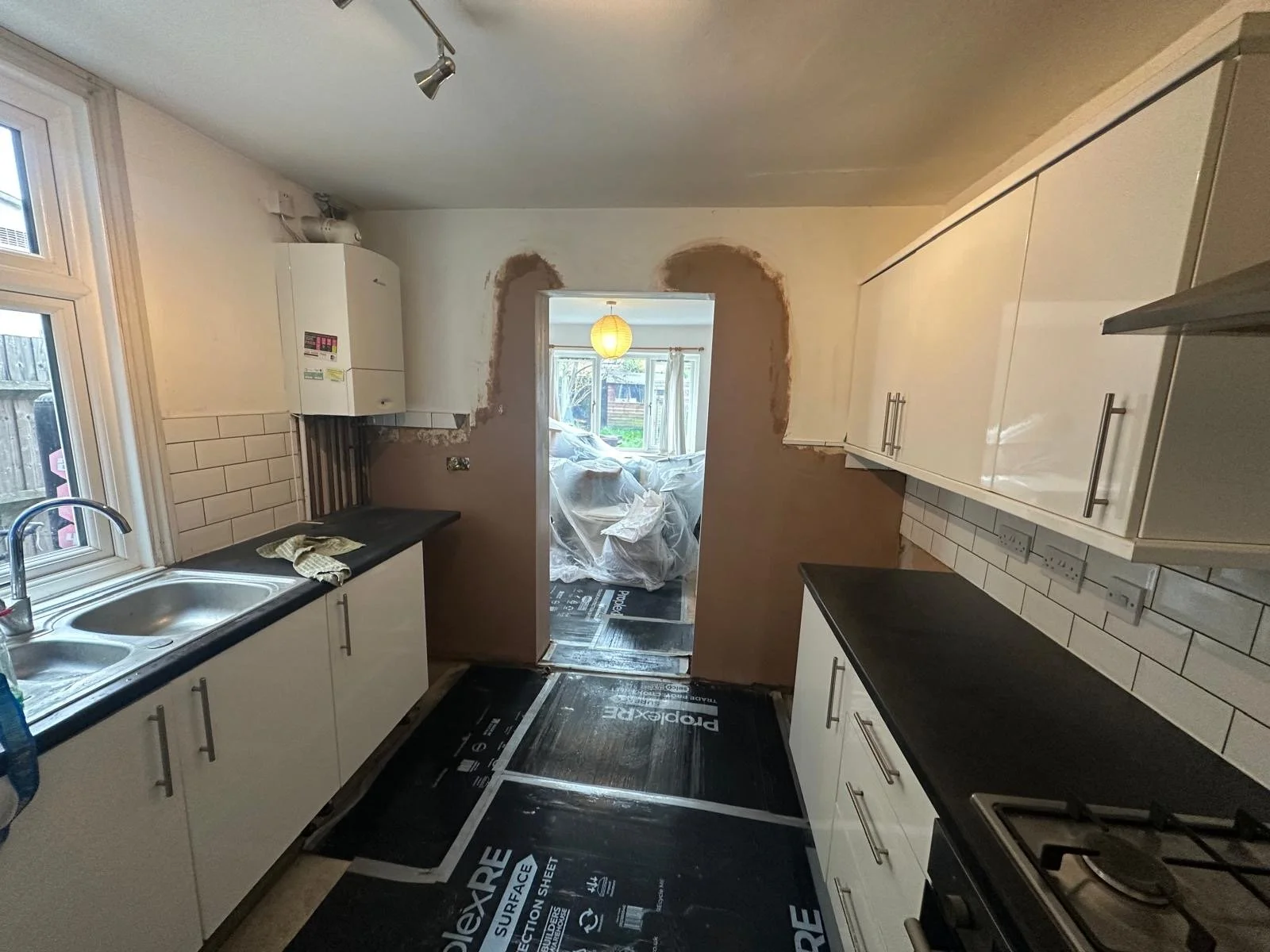 Kitchen with white cabinets, black countertop, stainless steel sink, white subway tile backsplash, window, and a doorway leading to another room with plastic-covered furniture and a hanging paper lantern.