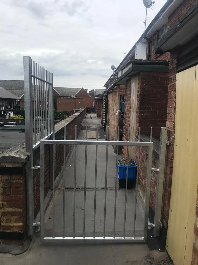 View of a narrow outdoor corridor with a metal gate at the entrance, brick walls on the sides, a blue plant pot on the right, and an overcast sky overhead.