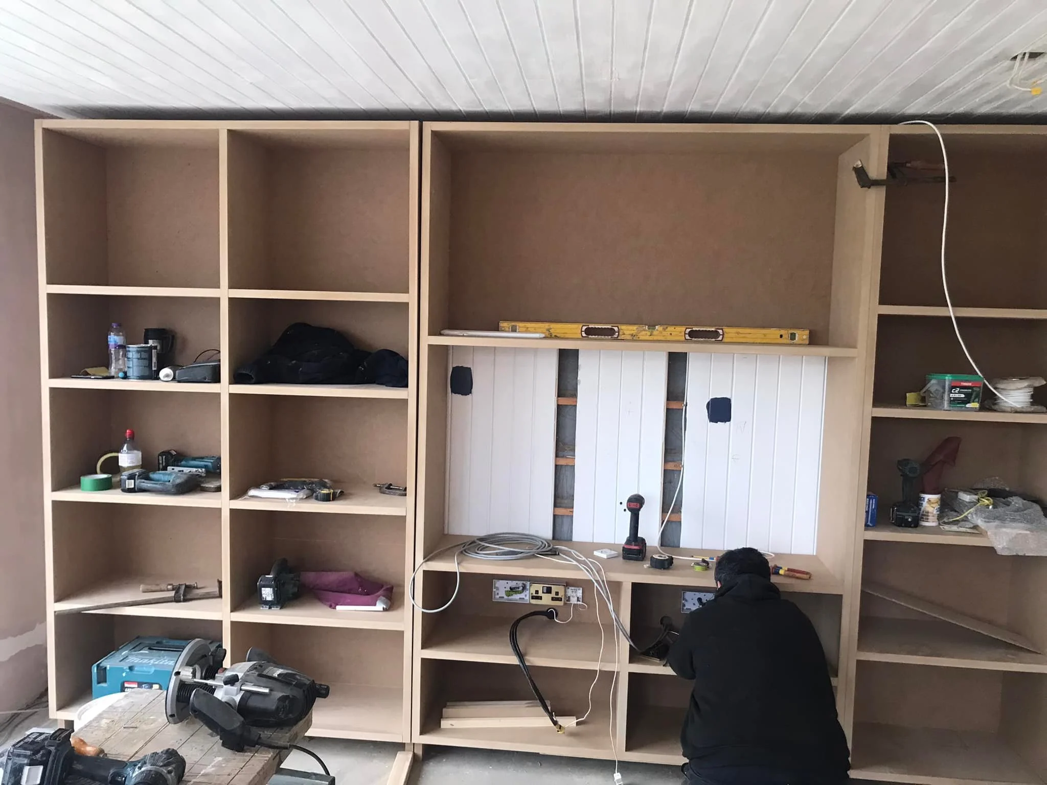 A worker installing cabinetry in a room, with tools and construction materials scattered around.