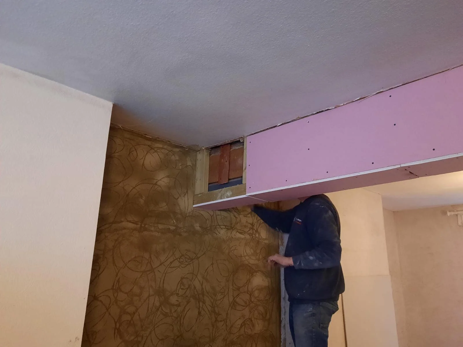 A person working for K G Croft working on drywall installation, standing on the floor with a partially finished wall and ceiling. There's pink drywall and exposed wood framing in north london.