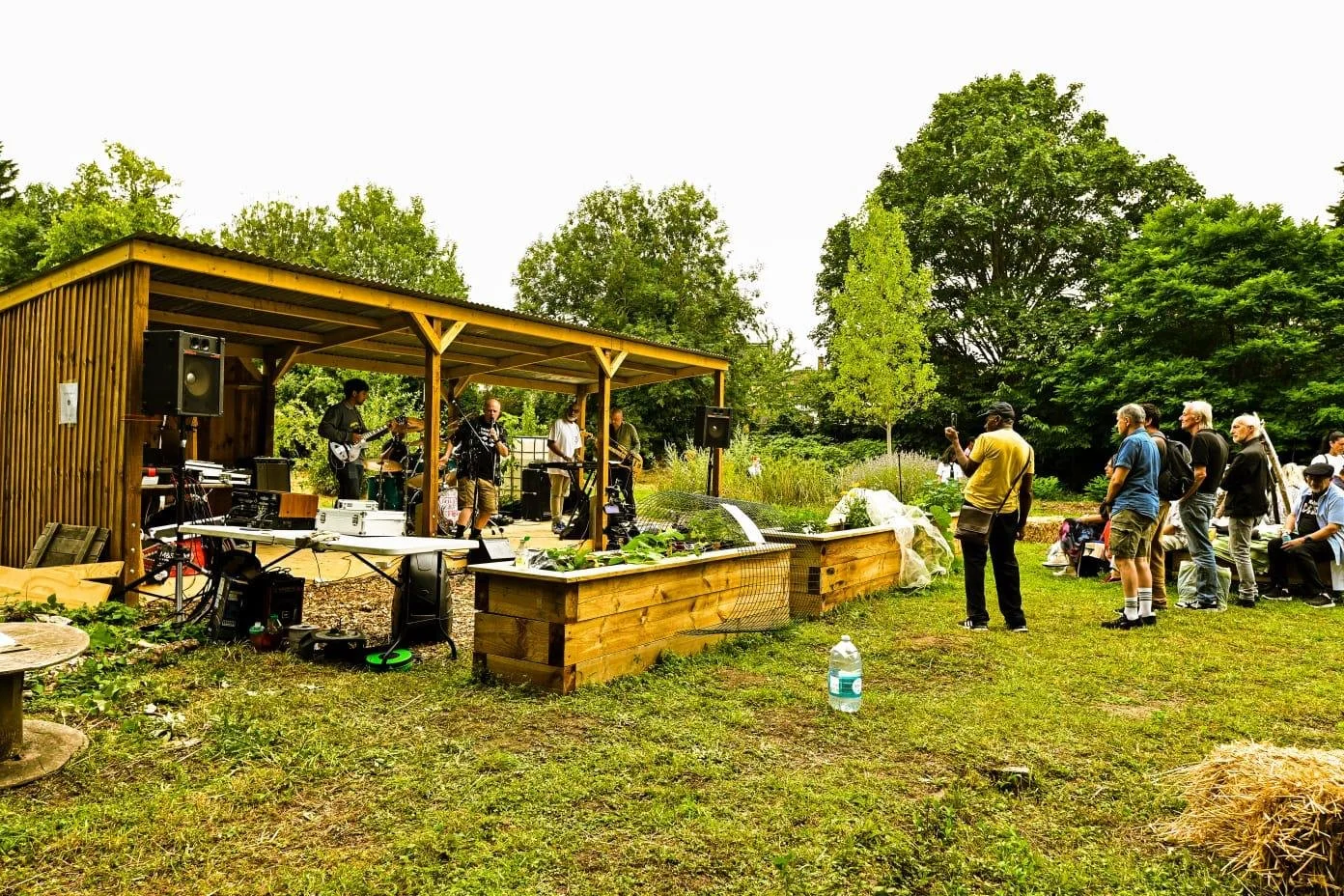 An outdoor concert in a park with a small band performing on a wooden stage, and audience members watching on a grassy area surrounded by trees. Arnos Park Sponsored by K G Croft.
