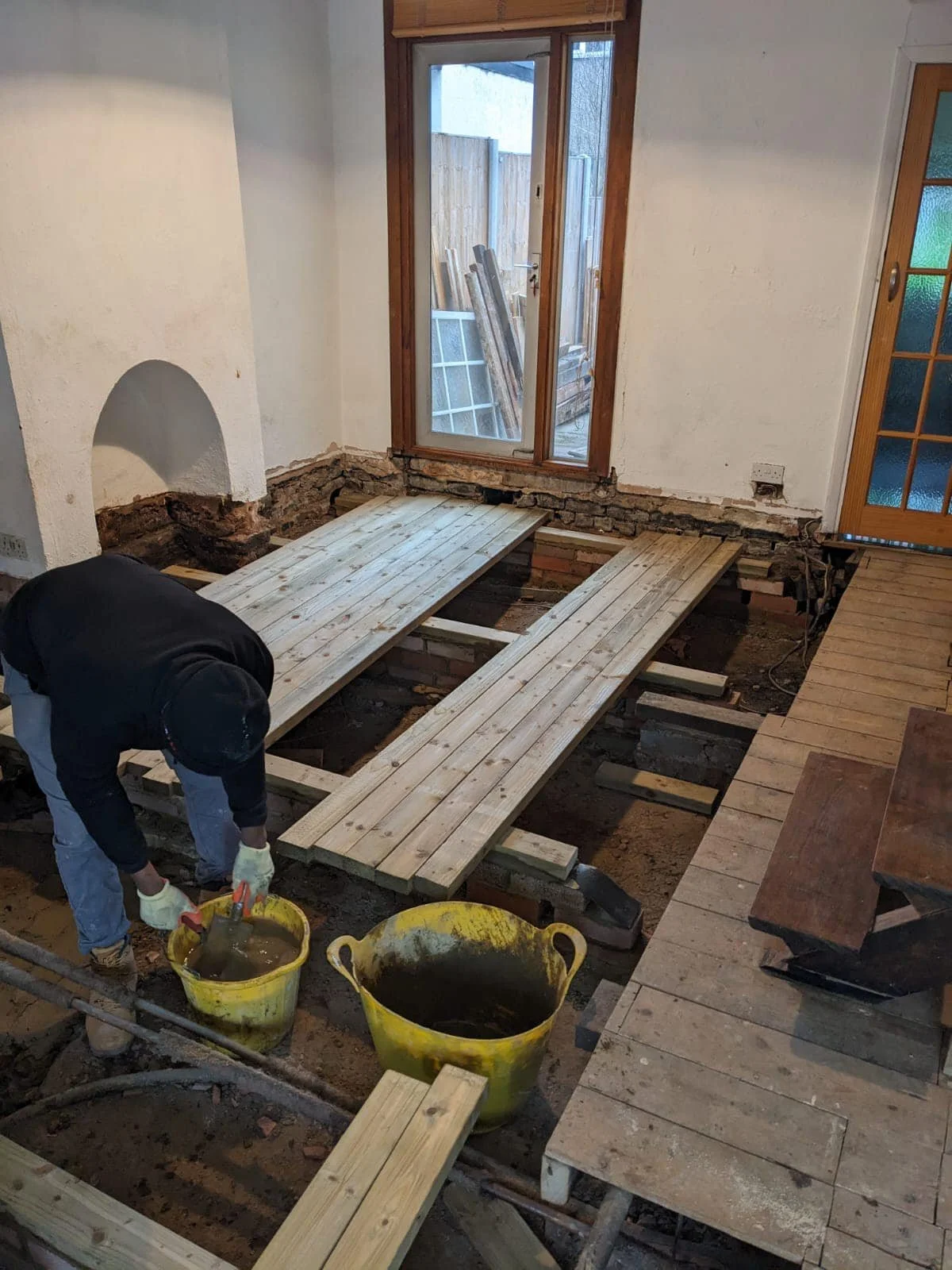 Person working on a floor renovation, installing wooden floorboards over a dugout area in a room with a glass door and a window for K G Croft in North London.