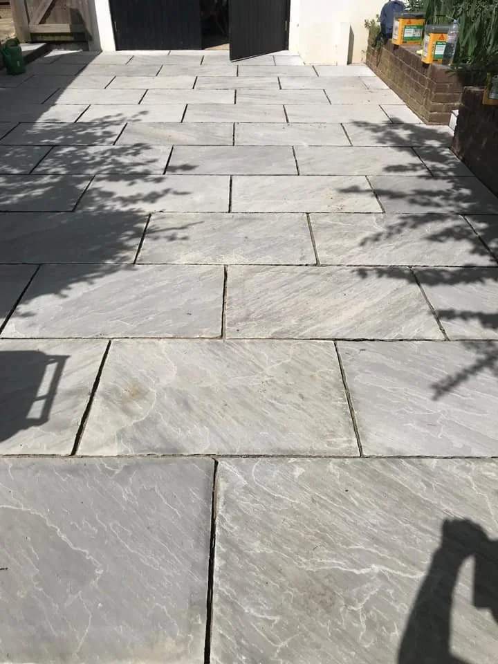 A freshly laid stone patio with large, rectangular, light gray stones arranged in a staggered pattern outside a building, with shadows of nearby plants and objects cast on it.