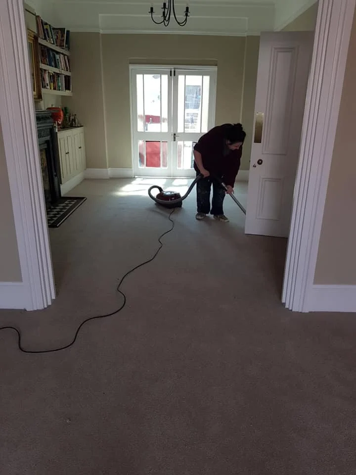 Person vacuuming a beige carpet in a living room with a window, bookshelf, and fireplace.