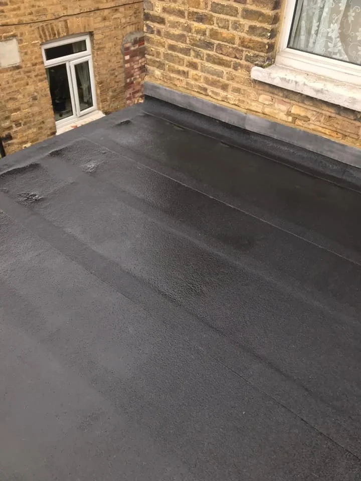 A rooftop in north london with fresh black waterproofing coating and a brick wall with a window in the background. Works by K G Croft.
