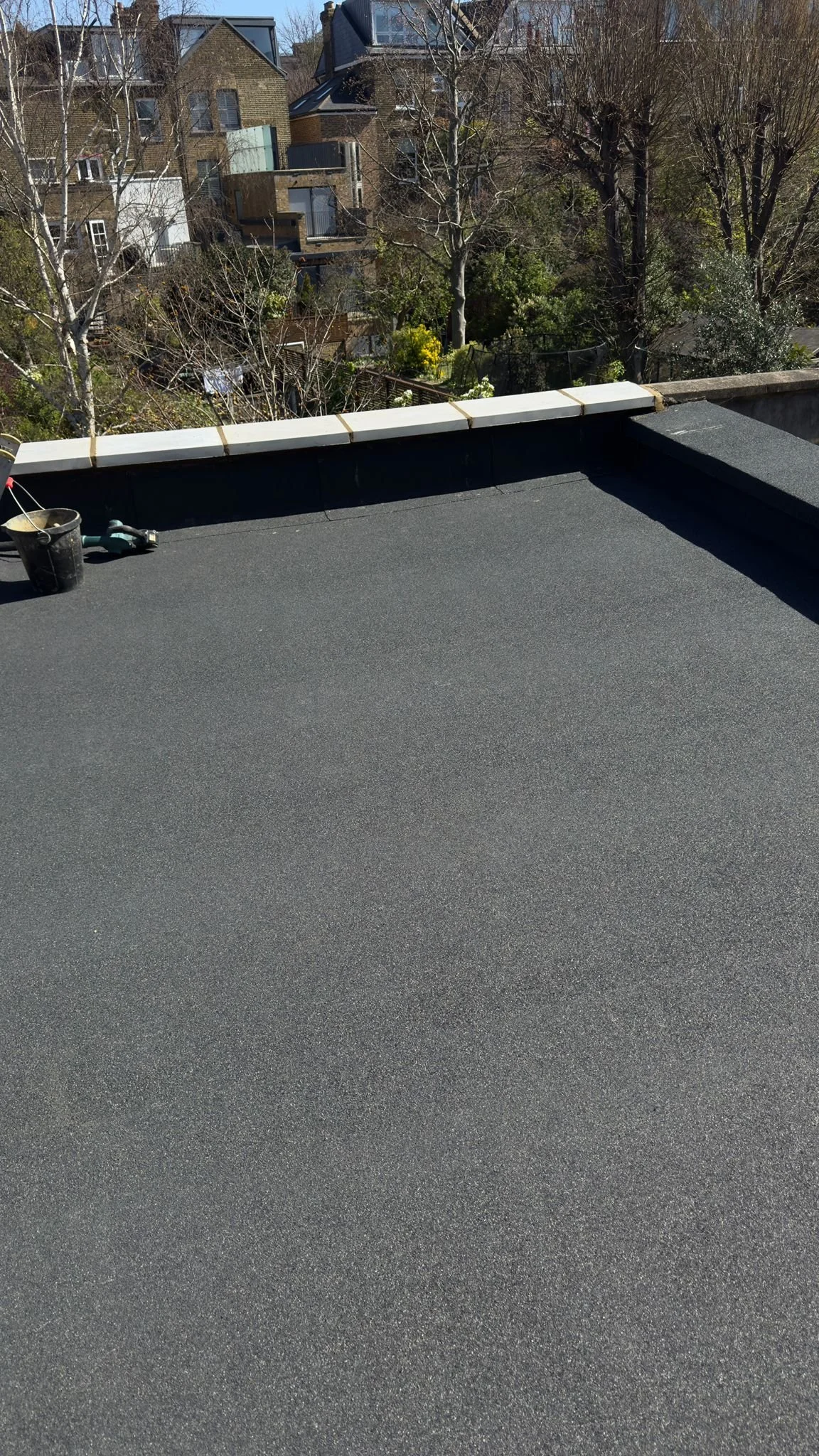 Roof with black asphalt shingles, tools and supplies on the edge, residential neighborhood with trees and houses in the background. Work by K G Croft in North London.