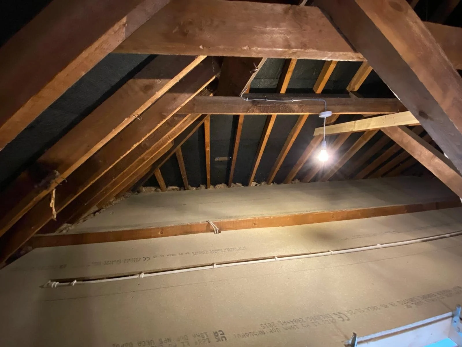 View of an attic ceiling with exposed wooden beams, insulation, and a hanging light bulb. K G Croft general building works.