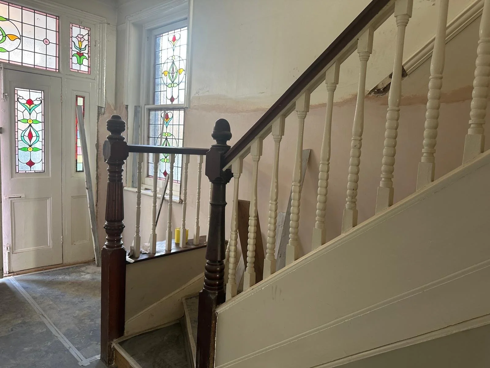 Interior of a house under renovation with stained glass front door, upstairs staircase, and a wall with peeling paint.