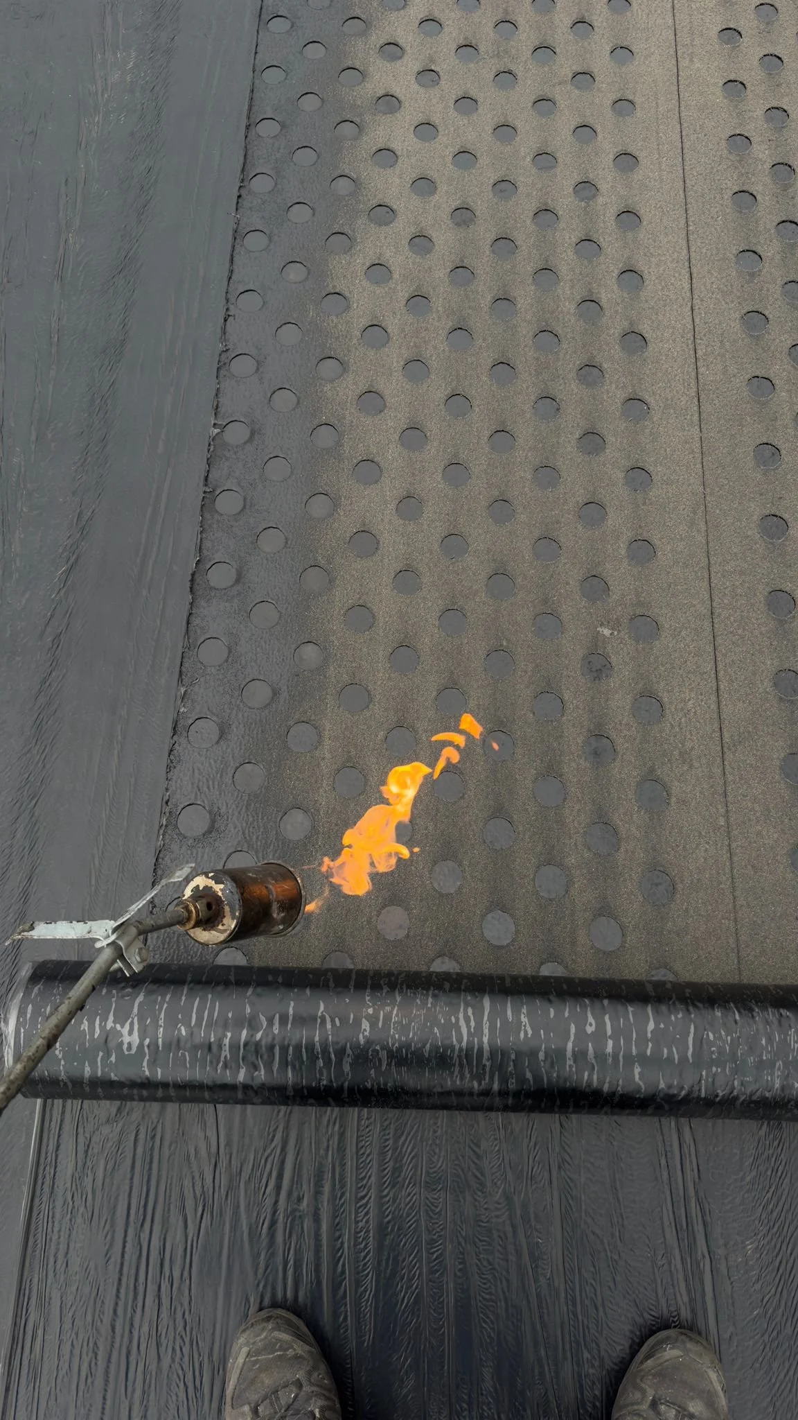 A person standing on a black textured surface near a doorway, using a torch to apply a flame to black adhesive or coating on a black pipe.