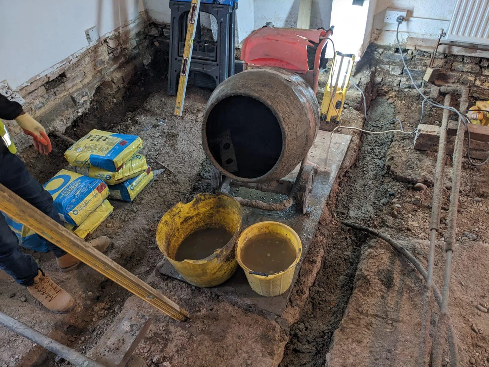 Construction site of K G Croft with an exposed foundation, a concrete mixer, buckets of mud, bags of cement, and tools for construction.