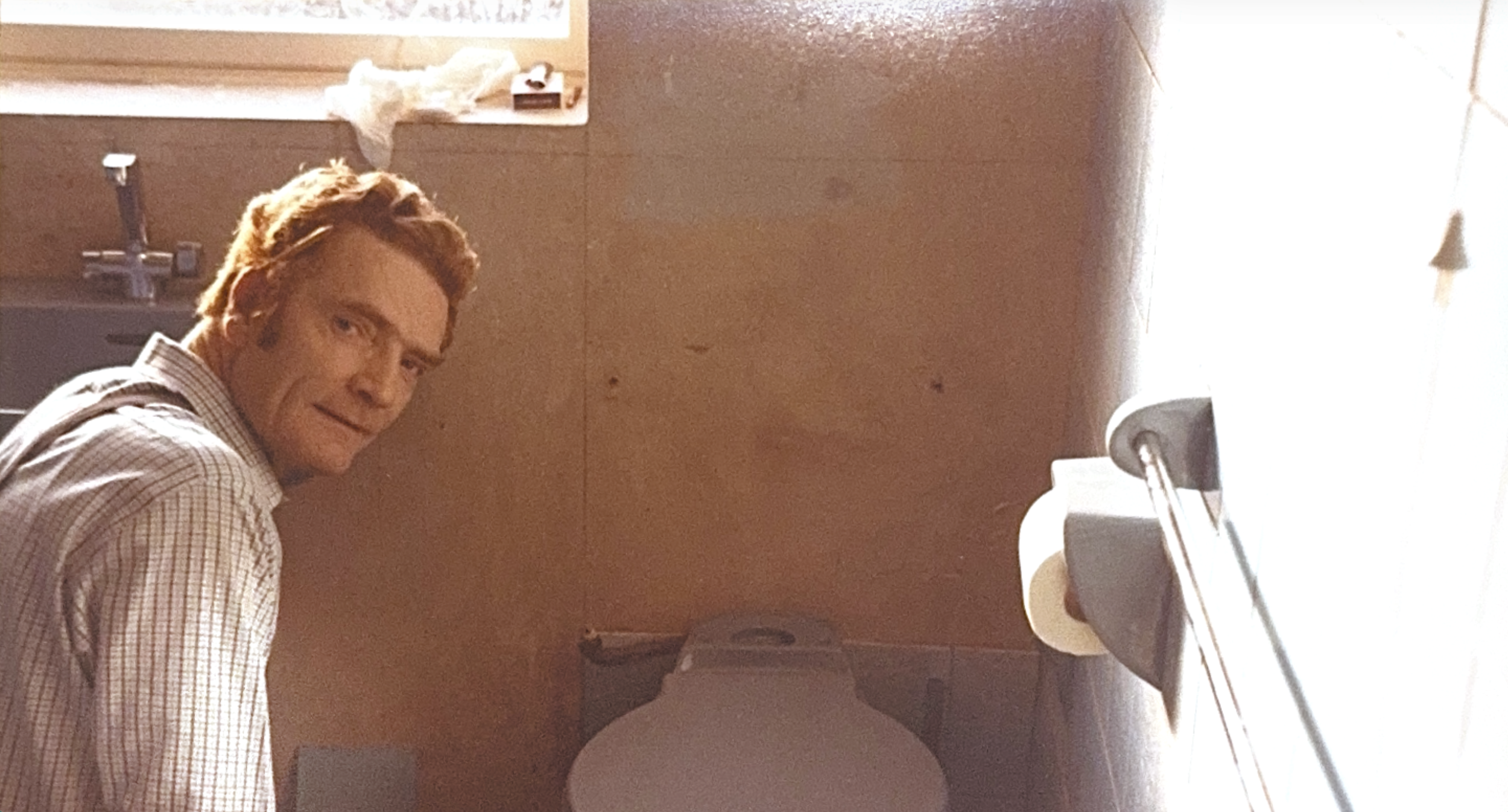 Kevin Croft's father with red hair and a checkered shirt inside a bathroom, sitting on the toilet, looking over his shoulder at the camera.