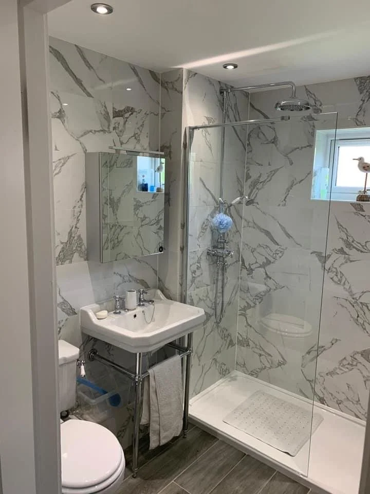 Small modern bathroom with marble-patterned walls, a white toilet, a small white sink with chrome legs, a mirror cabinet above, a glass walk-in shower with a rain shower head, a small window, and a gray tiled floor.