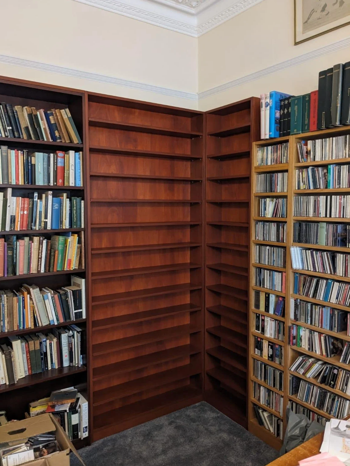 Wooden bookshelves built by K G Croft in a room in North London, with one partially filled with books and CD cases, and others mostly empty. There is a cardboard box and some items on the floor and a table in the foreground.