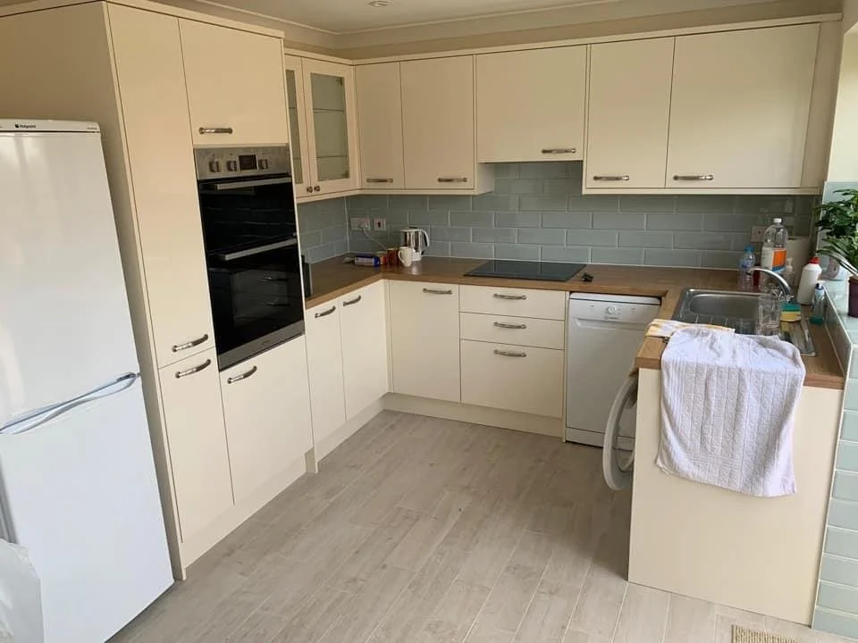 Kitchen with white cabinets, a refrigerator on the left, built-in oven, countertop with a kettle, sink with a towel, and various small appliances, including a water bottle and soap dispenser, on the right.