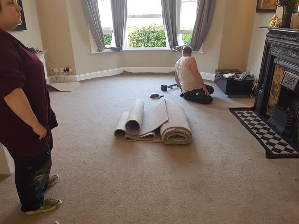 A person kneeling on a beige carpet near a large window, rolling out or installing a new carpet or rug, with another person standing and observing, in a living room with a decorated fireplace and window curtains.
