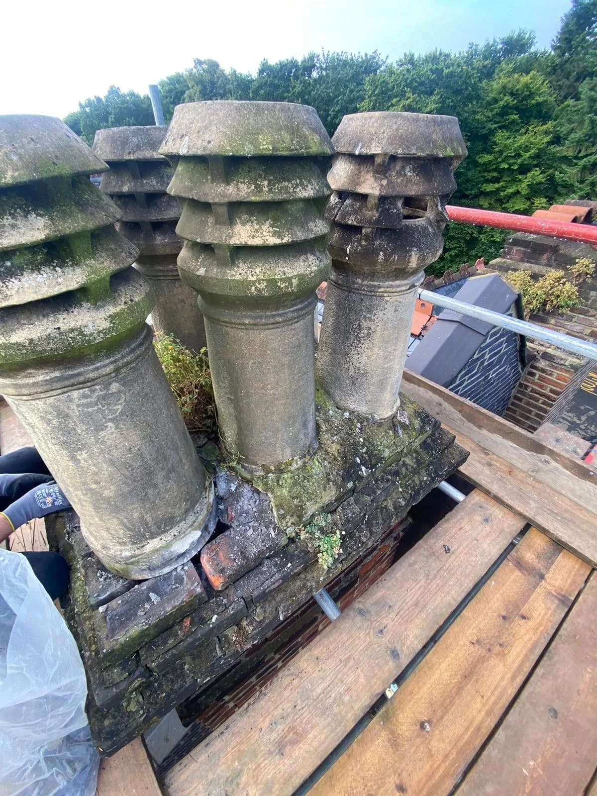 Close-up of moss-covered chimney pots on a rooftop, with scaffolding and trees in the background. Before roof works K G Croft.