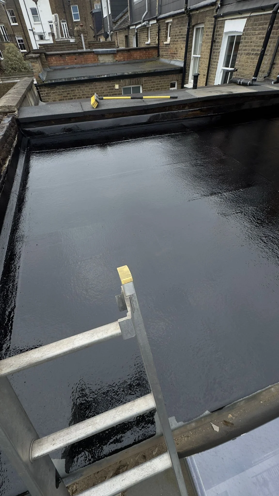 Roof surface being coated with black waterproof membrane, with a ladder and paint roller in use. North London work by K G Croft.