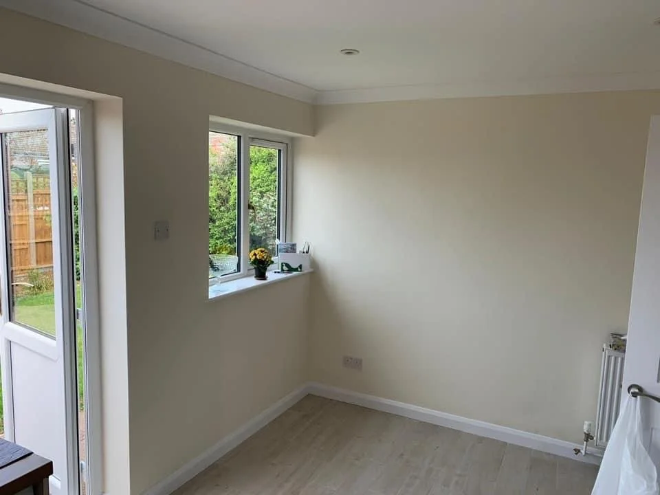 Empty room with white walls and light-colored flooring, adjacent to a door opening to a backyard with a wooden fence and grass, and a window with a small plant and decorative items on the windowsill.