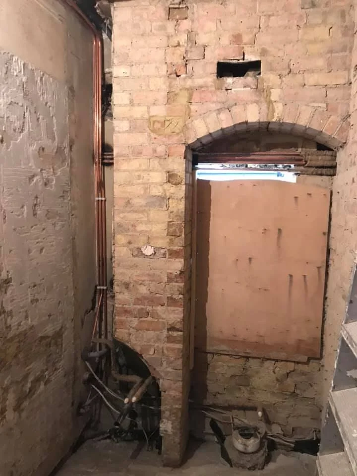 A brick wall with an arched window opening covered with a wooden plank, and exposed pipes and electrical wiring at the bottom corner in a construction or renovation site.
