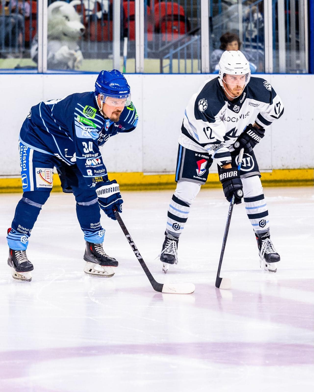 Two Harringay Huskies ice hockey players face off on the ice rink, preparing to compete for the puck, with a mascot dressed as a wolf visible behind the glass. Sponsored by K G Croft.