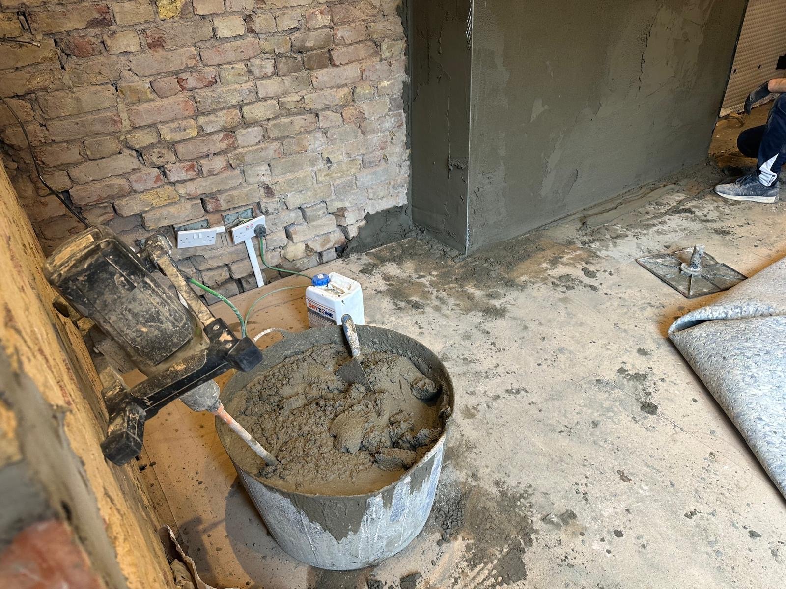 Construction site with a bucket of cement, a concrete mixer, an unfinished brick wall, an electrical outlet, and a person kneeling nearby.