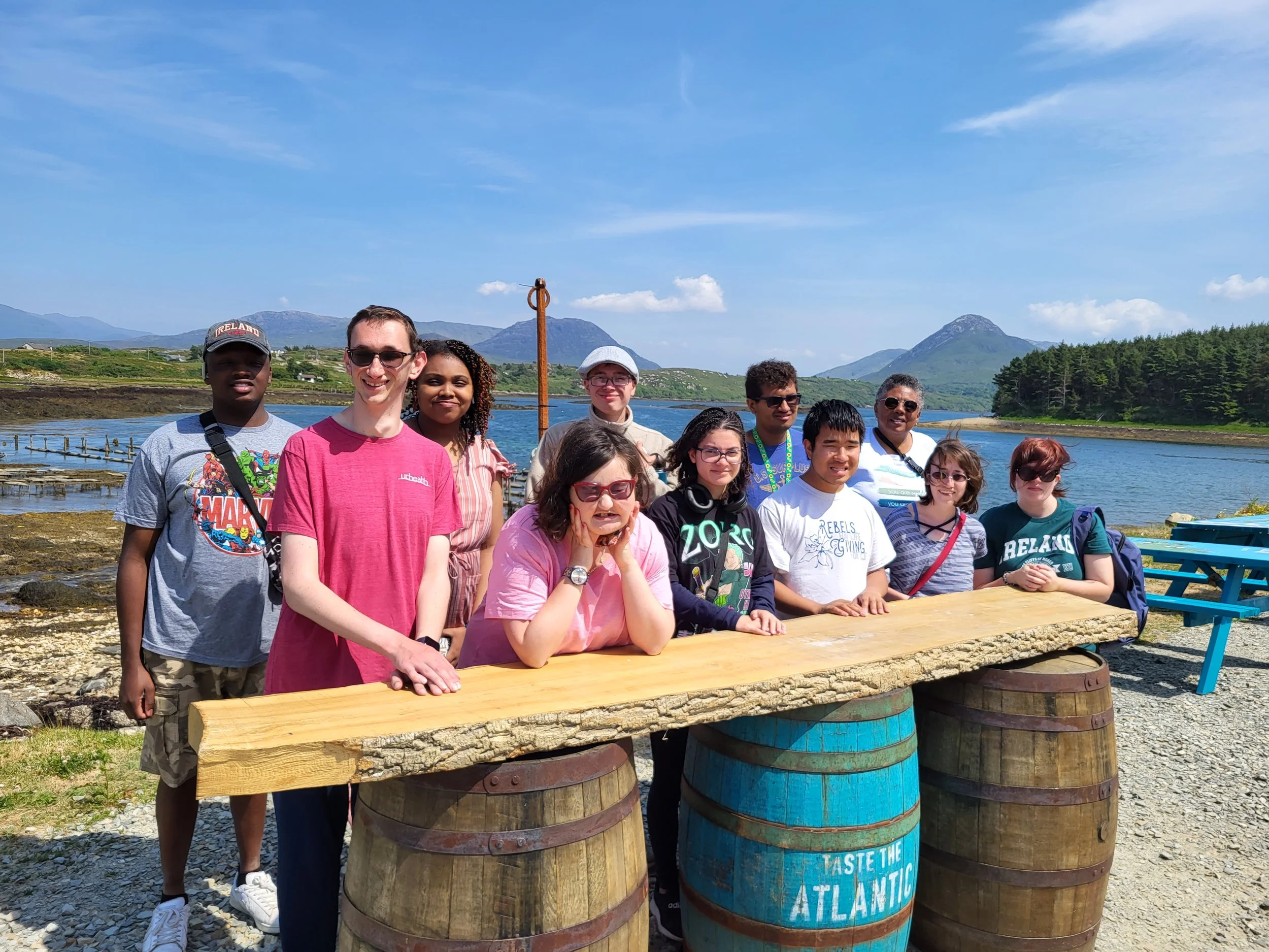 Group of young adults lean into a table made of barrels in front of a body of water- which is an oyster farm- in Ireland.