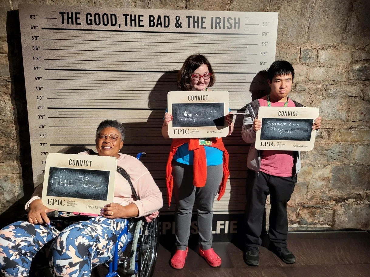 Group of three having fun in the immigration museum in Ireland. They stand in front of a sign that says, "the good, the bad, and the Irish". From left there is a woman in a wheelchair, who is holding up a sign that says "convict- The Pearl", she is s