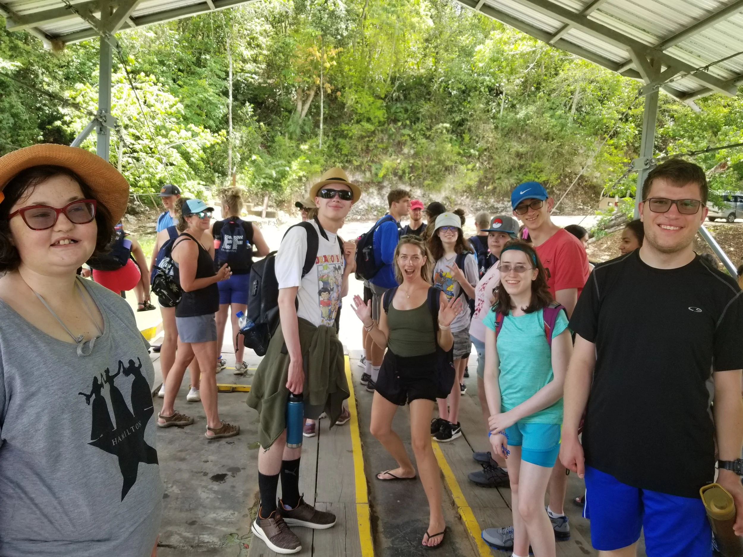 a group of travelers standing together smiling. Megan is in the middle doing a silly pose with a big "yay" face. Everyone is surrounded by green trees and water as we are on the barge to Xunantunich