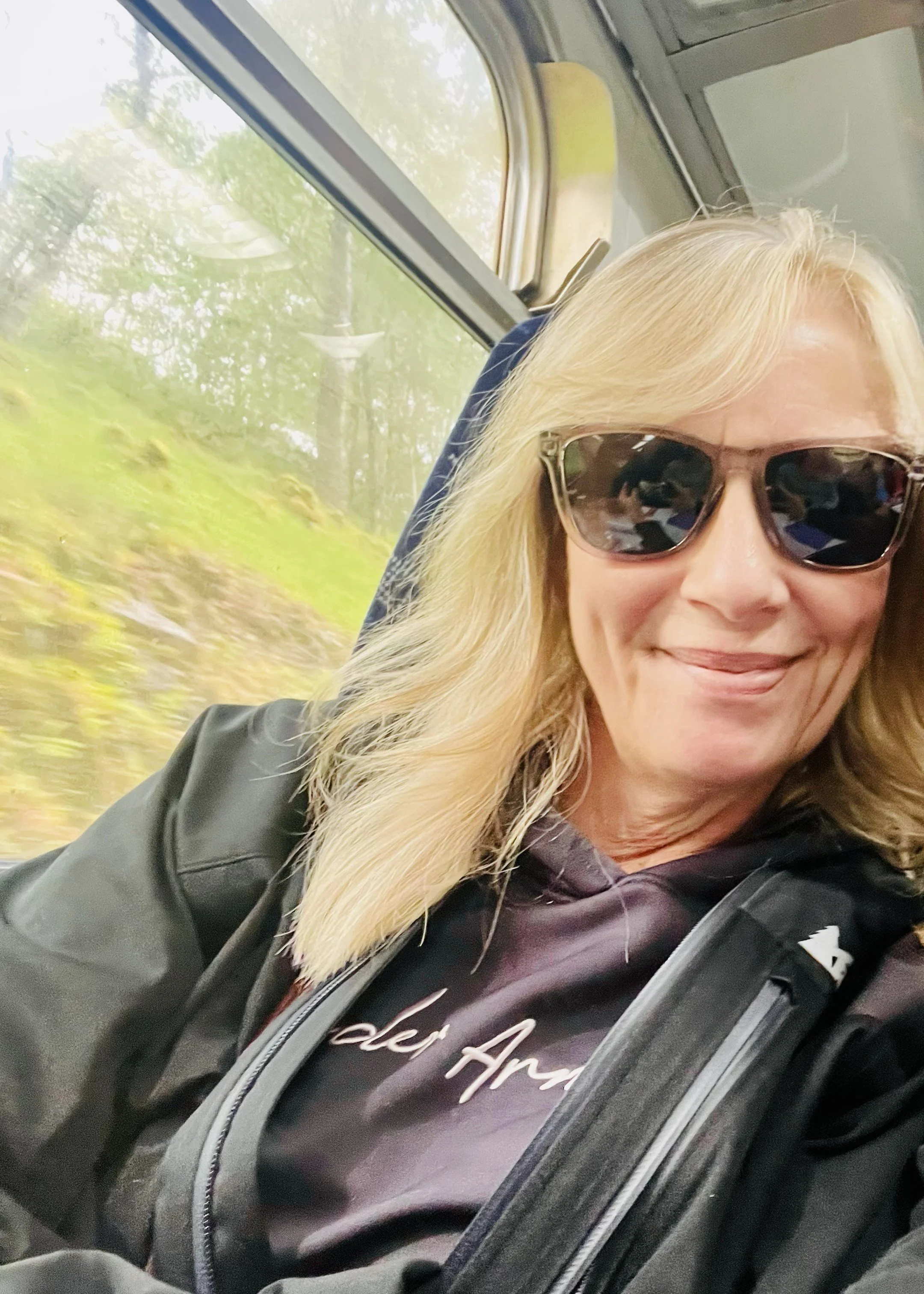 Ellen is smiling at the camera, wearing sunglasses and a black jacket with grey shirt. She has straight blonde hair down to her shoulders. Behind Ellen is a moving landscape, Ellen is on a moving train