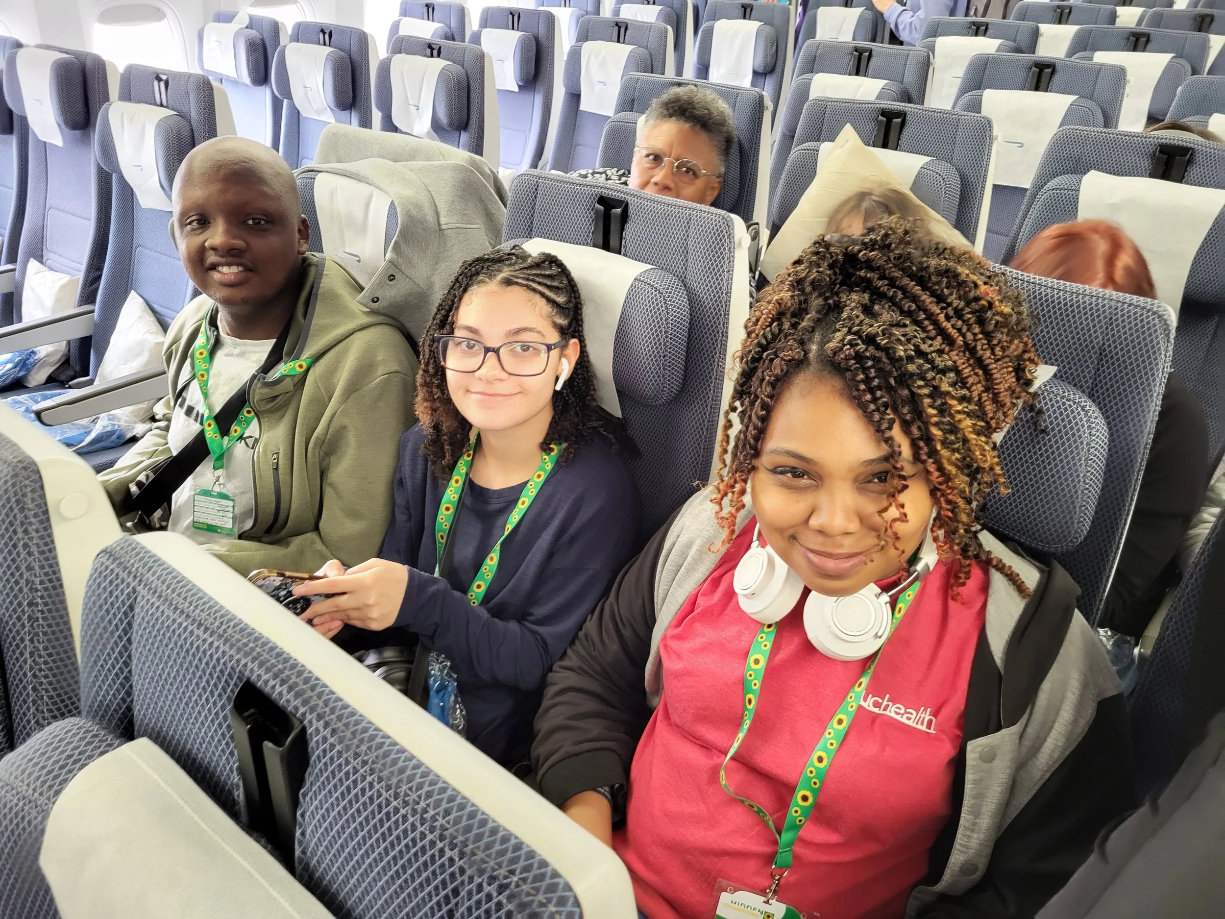 Three travelers, sitting in a plane, smile up at the camera from their seats.