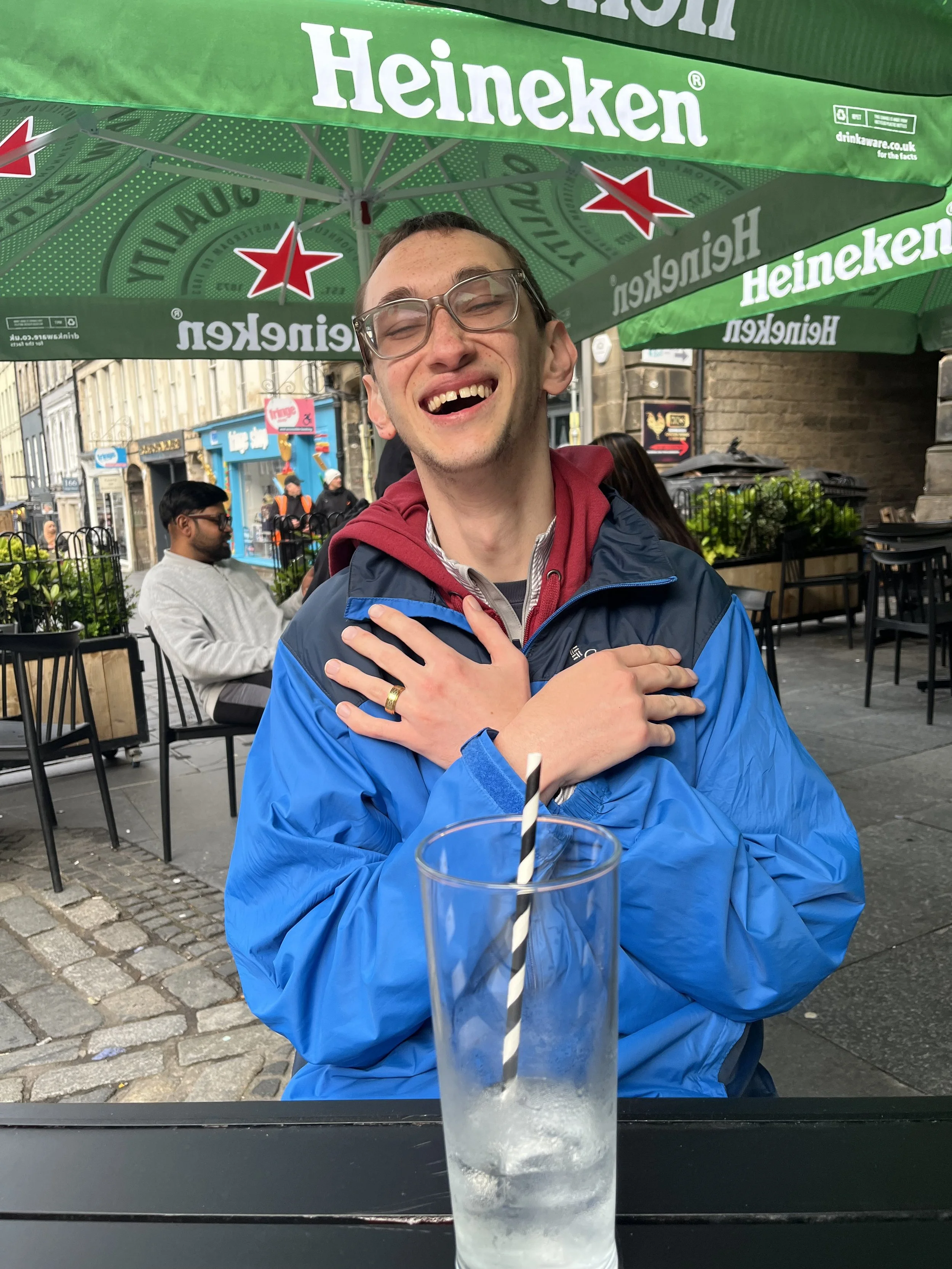 J.O.Y- A traveler is laughing, with his arms crossed in front of his chest. He is sitting at a table, with a water in front of him. The water has a straw that is black and white. 