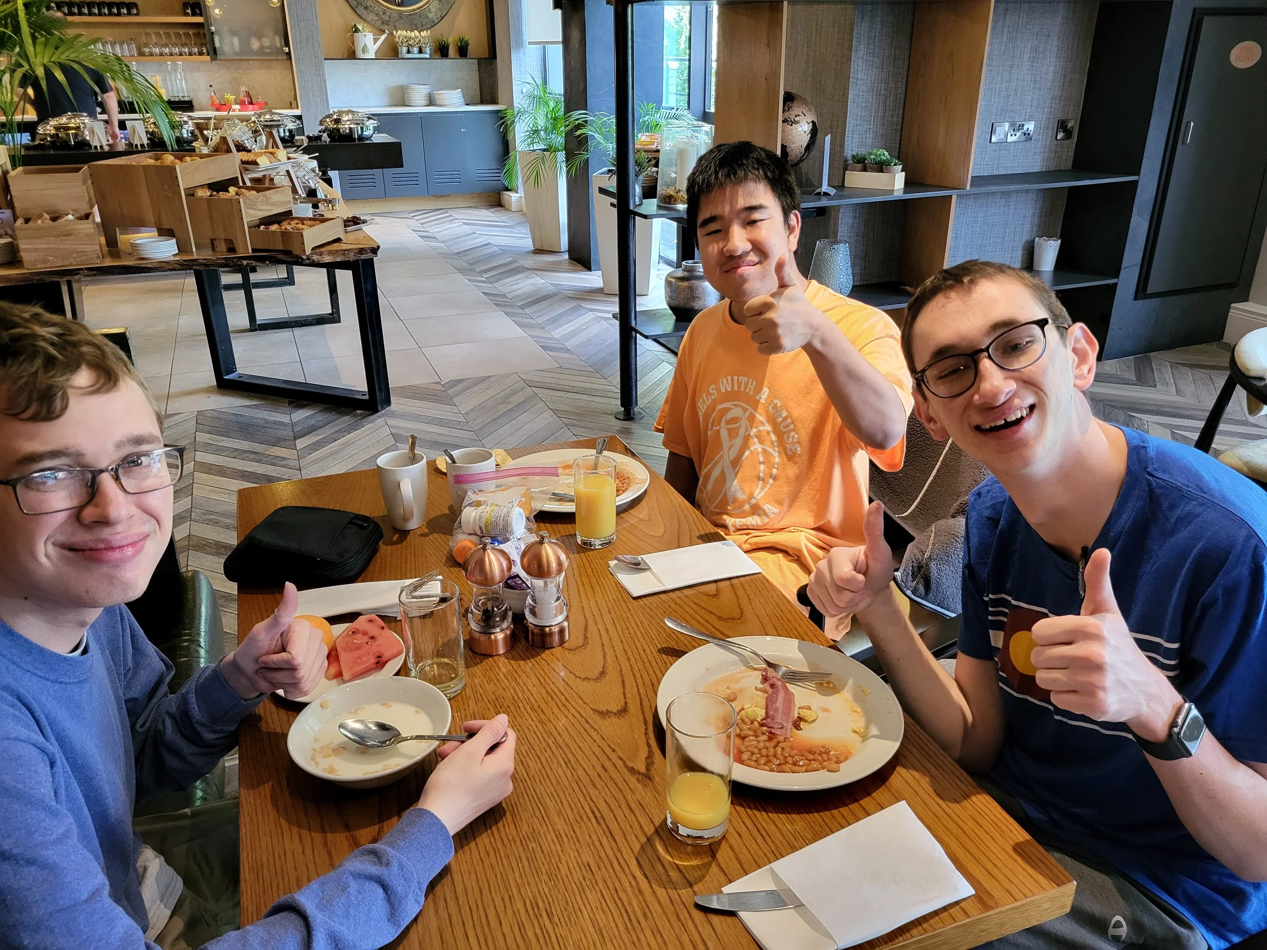Three young men, sitting at a table with breakfast items on their plates, looking at the camera, smiling with a thumbs up!